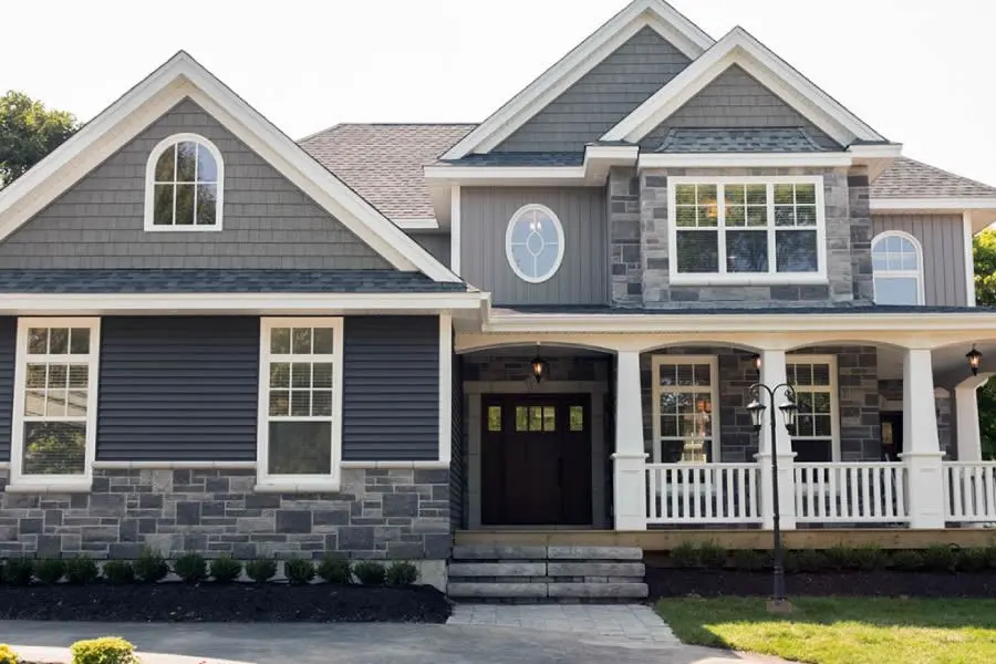 A stunning two-story home with designer siding featuring a mix of gray vertical board and batten, stone accents, and a charming front porch. This home showcases the expertise of a designer siding contractor i.e. Home Makeover.