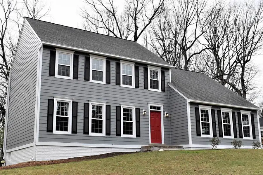 A two-story house with gray insulated vinyl siding, black shutters, and a red front door. The house has a gray roof and white trim around the windows and door. The house is surrounded by bare trees and a grassy lawn.