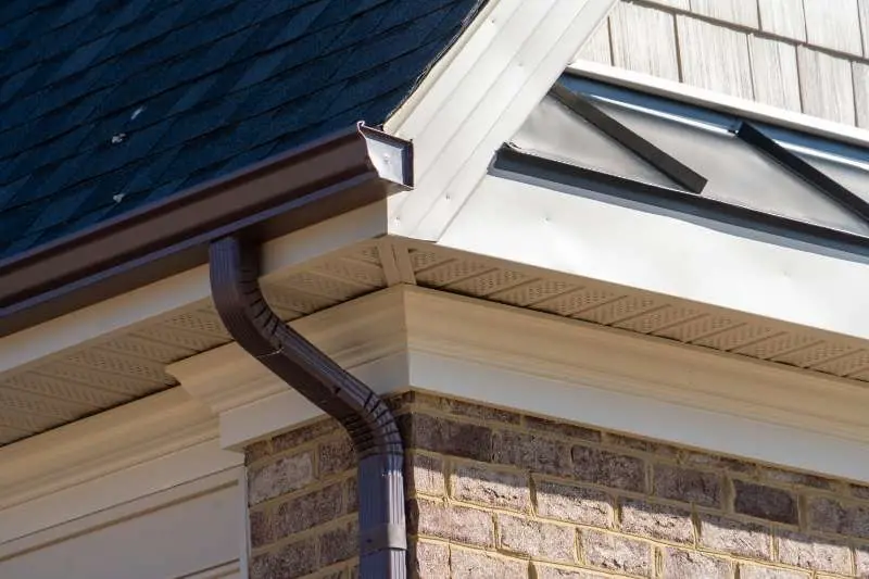 Close-up of a newly installed brown gutter and downspout on a brick house with a dark shingle roof, showcasing expert gutter replacement work.